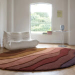 Rond Allegro High AEOH41891 vloerkleed met warme, golvende kleurbanen in een modern interieur met witte loungezetel.