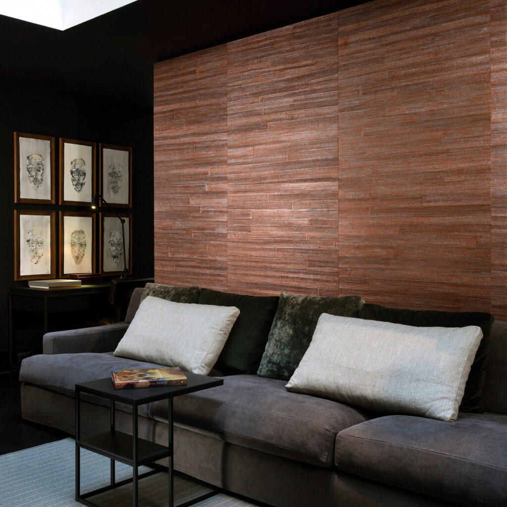 Sfeervolle woonkamer met Arte All Naturals Camago 14000 behang in warme bruintinten achter een donkergrijze bank met grote kussens.