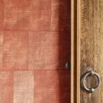 Rood Cuadro-behang uit de Gitane-collectie van Arte naast een houten deur met metalen ringgreep.