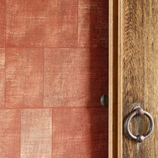 Rood Cuadro-behang uit de Gitane-collectie van Arte naast een houten deur met metalen ringgreep.