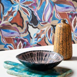 Decoratieve setting met Arte Essentials Tangram Myriad-24100 behang met kleurrijk bloemenmotief, een keramische schaal en een vaas op een tafel.