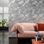 Moderne woonkamer met grijs Arte Facet behang van Buningh achter een lichtroze bank met oranje kussen.