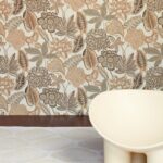 Bloemrijk Arte Fiori Antichi 49560 behang uit de Gitane collectie met warme aardetinten, naast een lichtgekleurde designstoel.