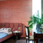 Sfeervolle slaapkamer met rood Arte Fringe behang, fluwelen bed, houten bureau en grote groene kamerplant.
