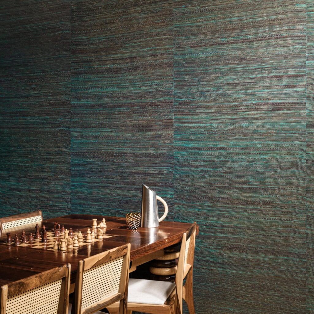 Houten tafel met schaakbord en stoelen tegen een muur bekleed met Arte Icarus Penumbra 15741 wandbekleding in blauwgroene tinten.