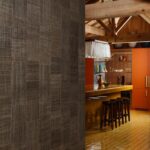 Moderne keuken met een opvallende okergele bar, oranje kasten en een muur bekleed met Arte Icarus Telaio 15729 wandbekleding in een warme, geweven textuur.