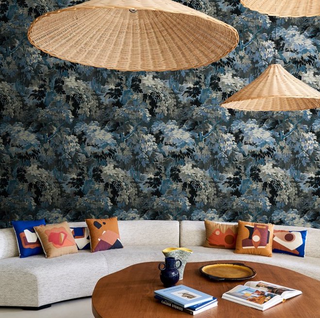 Stijlvol interieur met Arte Les Forêts Diore 48013 behang in cobalt-blauw, rotan hanglampen en kleurrijke kussens op een beige bank.