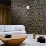 Modern interieur met een lichtgrijze bank en houten tafel tegen een decoratieve zwarte muur met goud patroon van Arte Joie 30811 behang.