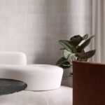 Moderne zithoek met Arte KatanSilk 11505 wit behang, een ronde witte sofa, een plant en een marmeren salontafel.