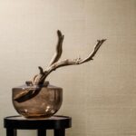 Glazen vaas met decoratief stuk hout staat voor beige Arte Line behang 80700B.