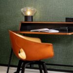 Designbureau met een leren Gioco-stoel van Arte en een tafellamp tegen een groene linnenstructuur wandbekleding.