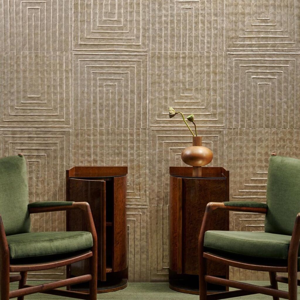 Twee groene fauteuils en houten bijzettafels staan voor een wand bekleed met Arte Luster Cornice 70332 met geometrisch reliëfpatroon.