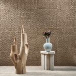 Moderne interieursetting met Arte Luster Perula 70302 wandbekleding, een houten sculptuur en een vaas met takken op een houten tafeltje.