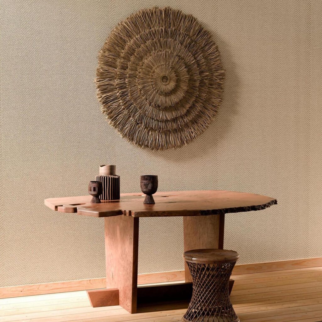 Houten tafel met decoratieve objecten, een gevlochten kruk en een wanddecoratie in natuurlijke stijl tegen behang uit de Arte Marquesa Atuona 31721 collectie.