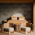 Slaapkamer met een tropisch geïnspireerd interieur, inclusief een bed met okergeel beddengoed, twee stoelen met natuurlijke materialen en een Arte Marquesa Fenua wandbekleding met palmbladmotief.