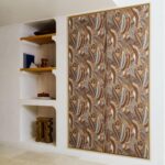 In een modern interieur hangt het wandpaneel Arte Melaky Mandrava 40761 met een organisch bruin en okergeel patroon naast een ingebouwde witte wandkast met houten planken.