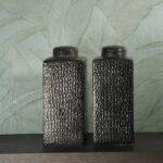 Twee zwarte, rechthoekige vazen met krokodillenstructuur staan voor een groene muurbekleding met groot bladmotief uit de Arte Monsoon Musa collectie.