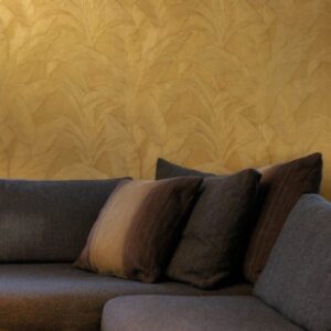 Donkergrijze bank met kussens tegen een goudkleurige Arte Monsoon Musa wand met bladmotief.