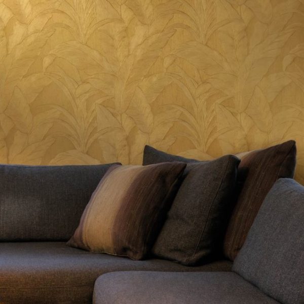 Donkergrijze bank met kussens tegen een goudkleurige Arte Monsoon Musa wand met bladmotief.