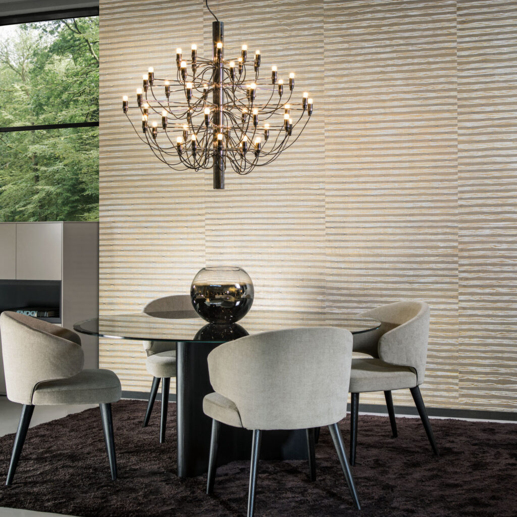 Moderne woonkamer met Arte Raphia behang, een ronde glazen eettafel, vier beige stoelen en een opvallende zwarte kroonluchter.