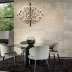 Moderne woonkamer met Arte Raphia behang, een ronde glazen eettafel, vier beige stoelen en een opvallende zwarte kroonluchter.