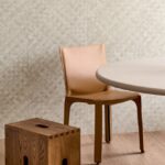 Moderne interieurhoek met Arte Textura Pandan-behang in zandkleur, een leren stoel, houten kruk en ronde tafel.