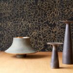 Decoratief Arte Ajoura Ebony Les Forêts behang met gouden bloemmotief, gecombineerd met keramische designobjecten op een houten oppervlak.