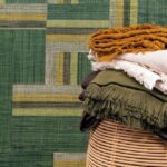 Stapel kleurrijke stoffen op een rotan mand met op de achtergrond het geometrische Arte behang Atelier Boro AT71 in groen- en geeltinten.