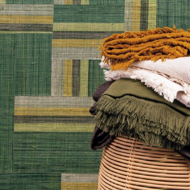 Stapel kleurrijke stoffen op een rotan mand met op de achtergrond het geometrische Arte behang Atelier Boro AT71 in groen- en geeltinten.