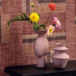 Twee roze keramische vazen met bloemen staan voor een roodbruin Arte Atelier Boro behang met textuurpatroon.