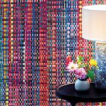 Kleurrijk Arte Atelier Extravaganza behang met geweven patroon achter een tafeltje met bloemenvaas en lamp.