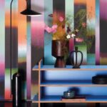 Modern interieur met Arte-behang Atelier Time Lapse in kleurrijke verticale strepen, gecombineerd met een blauwe wandkast, bloemenvaas en designlamp.