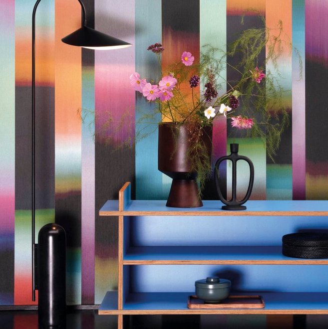 Modern interieur met Arte-behang Atelier Time Lapse in kleurrijke verticale strepen, gecombineerd met een blauwe wandkast, bloemenvaas en designlamp.