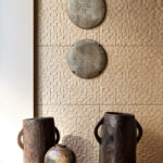Decoratieve wand met Arte-behang uit de Babylon-collectie in de kleur Adobe, gecombineerd met houten vazen en ronde wandornamenten.