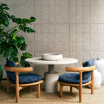 Moderne zithoek met houten stoelen en ronde tafel tegen een Arte Babylon Terracotta 97012 behangwand met reliëfpatroon.