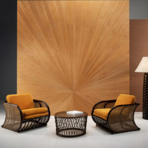 Twee rotan loungefauteuils met okergele kussens en een ronde rotan salontafel staan voor het Arte-behang Boutique Sunburst 90511 met een opvallend zonnestraalpatroon.