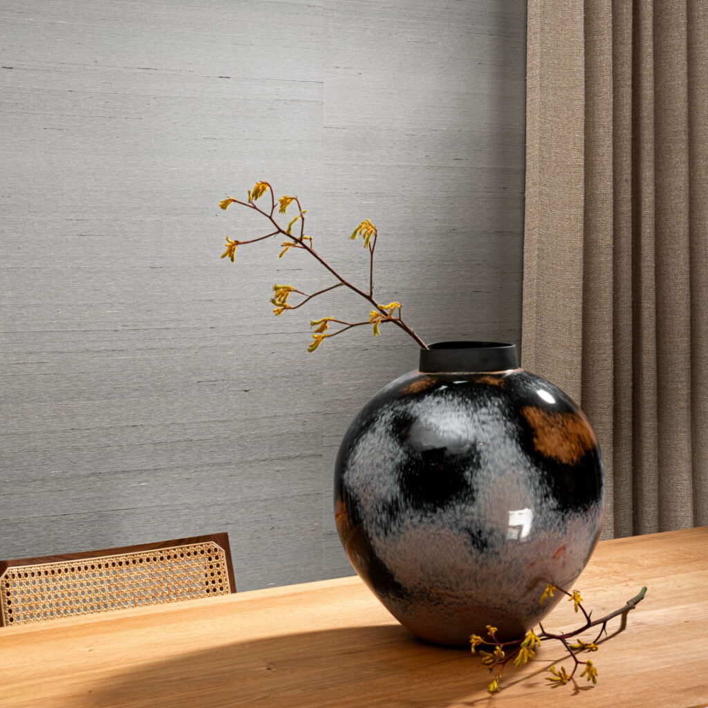 Grote bolvormige vaas met een takje gele bloemen op een houten tafel, voor Arte Dupion behang in een sfeervol interieur.