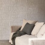 Beige zitbank met neutrale kussens en plaid staat voor een muur met Arte Essentials Costura Bojagi 57542 behang in een subtiel geometrisch patroon.