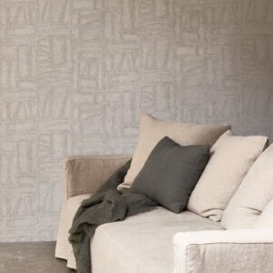 Beige zitbank met neutrale kussens en plaid staat voor een muur met Arte Essentials Costura Bojagi 57542 behang in een subtiel geometrisch patroon.
