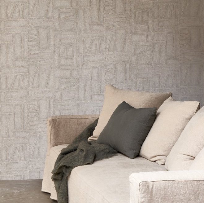 Beige zitbank met neutrale kussens en plaid staat voor een muur met Arte Essentials Costura Bojagi 57542 behang in een subtiel geometrisch patroon.