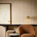 Interieur met Arte-behang Essentials Les Naturels Le Raphia Tissé 26746, een warme stofstructuur op de wand, gecombineerd met een bruine fauteuil, gouden tafellamp en abstract kunstwerk.