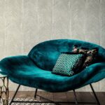 Fluwelen turquoise fauteuil met decoratieve kussens voor een muur met Arte-behang Essentials Talamanca in een subtiel patroon.