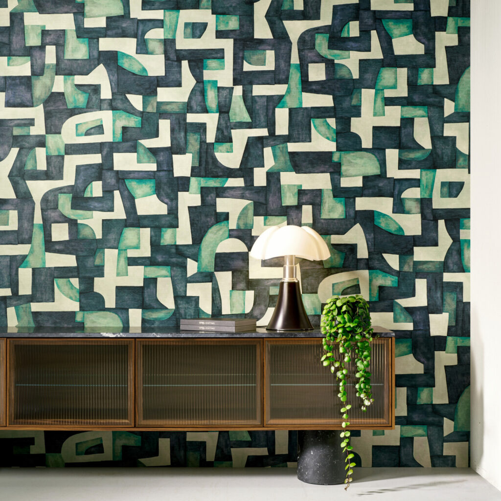 Interieur met Arte behang Essentials Totem Ecoline 18901, een abstract geometrisch patroon in groen- en beige-tinten, achter een retro dressoir met lamp en hangplant.