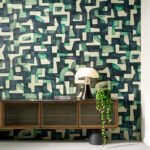 Interieur met Arte behang Essentials Totem Ecoline 18901, een abstract geometrisch patroon in groen- en beige-tinten, achter een retro dressoir met lamp en hangplant.