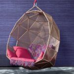 Hangstoel met roze kussens en franjerand, voor een muur met paars Arte-behang uit de Expedition Tasar collectie.