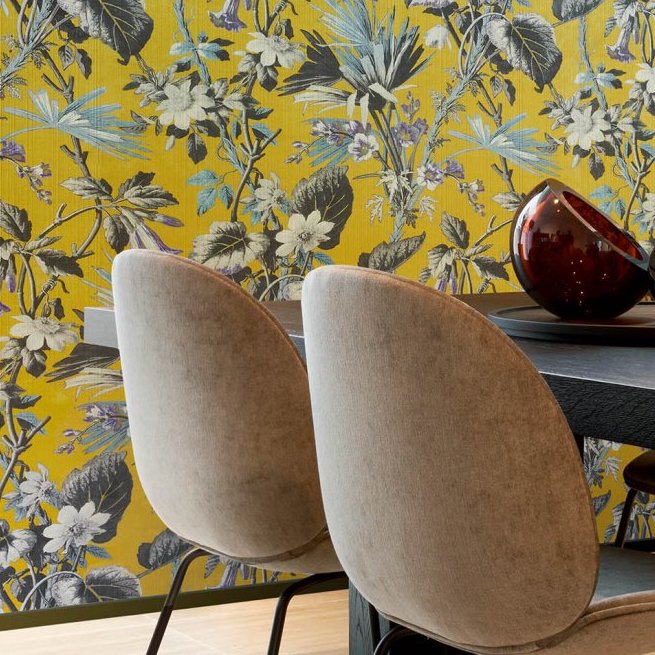 Twee beige stoelen aan een zwarte tafel met op de achtergrond geel Arte-behang Insolence Grow met een weelderig bloemenpatroon.