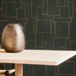 Arte-behang Insolence Nerve met goudkleurig lijnenspel op donker oppervlak achter een licht houten tafel met een metallic vaas.