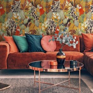 Interieur met een roestkleurige fluwelen bank, kleurrijke kussens, een ronde salontafel en het opvallende Arte-behang JV800 Tierra Giungla met tropisch junglepatroon.