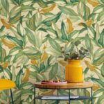 Sfeervolle interieursetting met het Arte behang JV800 Tierra Mediterraneo 6611 met groen-geel bladmotief, een gele vaas met takken en een houten tafel.