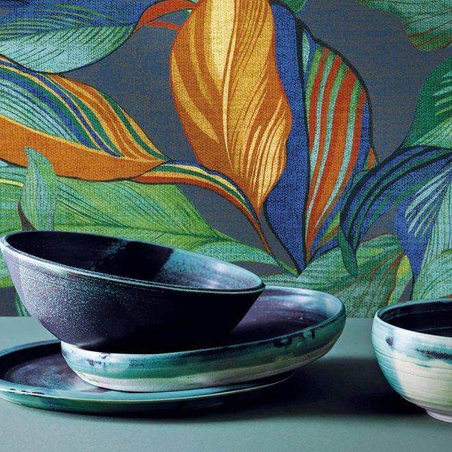 Keramische kommen en borden in aardetinten staan voor een wand met kleurrijk Arte Tierra Mediterraneo behang met tropisch bladmotief.